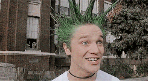 A punk with green spikey hair and bright orange gloves on pretends to be scared in a clearly sarcastic way